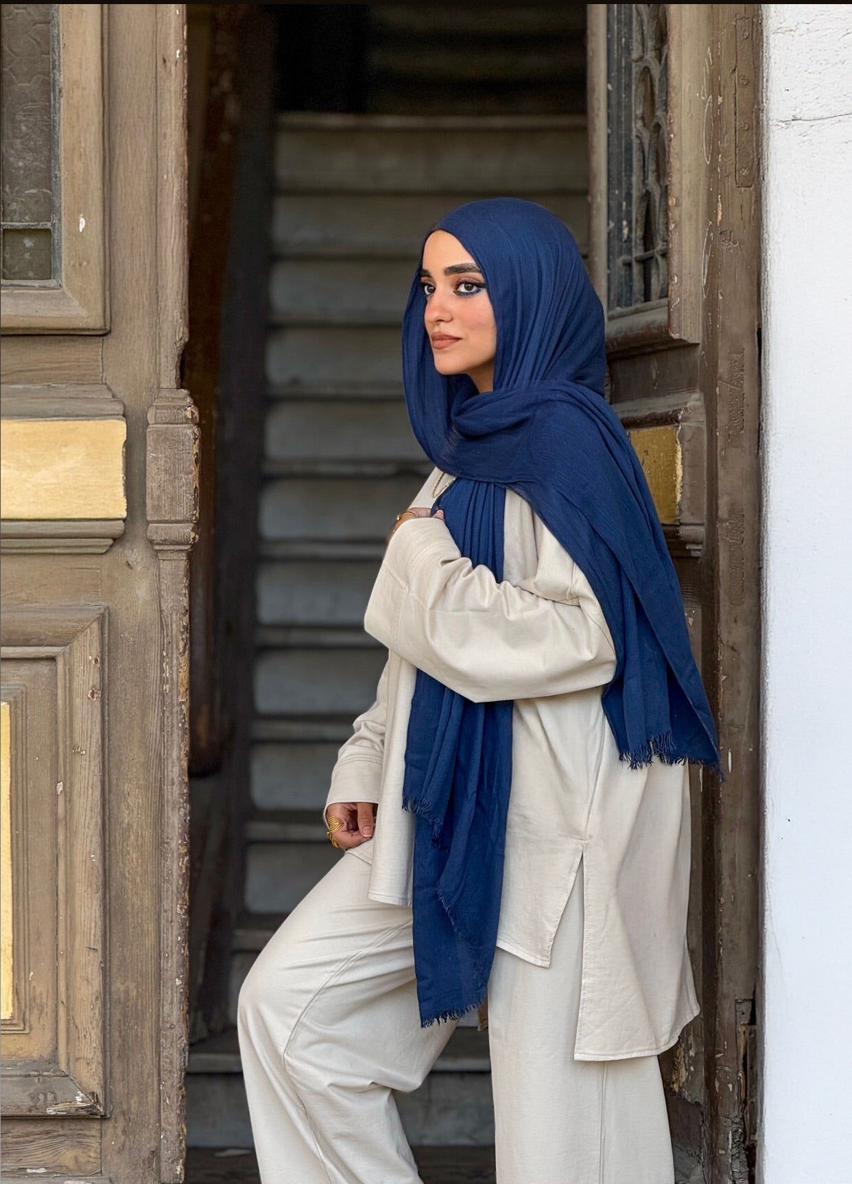 Woman wearing a blue hijab and white outfit standing in front of a wooden door.